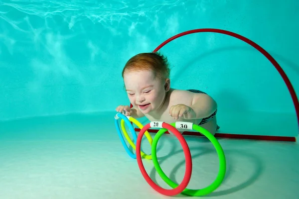Un niño feliz con síndrome de Down nada y recoge juguetes bajo el agua del fondo de una piscina infantil durante una actividad deportiva. Discapacidad infantil. Concepto. Retrato. Orientación horizontal. — Foto de Stock