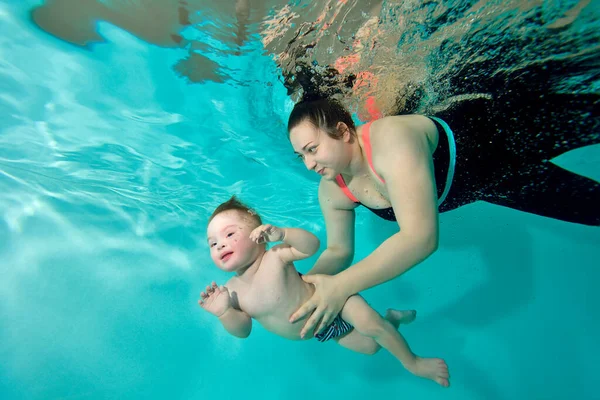 Una madre feliz y un niño con síndrome de Down nadan juntos bajo el agua en una piscina de agua turquesa. La mujer sostiene al bebé con sus manos y sonríe. Retrato. Orientación horizontal. — Foto de Stock