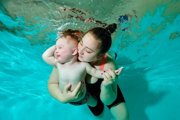 Una madre feliz abraza firmemente a su bebé con síndrome de Down y nada con él bajo el agua en una piscina infantil con agua turquesa. Discapacidad infantil. Concepto. Retrato. Orientación horizontal. Imagen De Stock