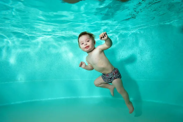 Un niño, un niño pequeño, con síndrome de Down nada bajo el agua y posa en una piscina con agua turquesa. Orientación horizontal. Imagen De Stock