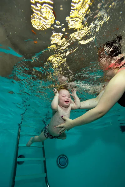 Щаслива дитина з синдромом Дауна плаває до рук своїх матерів під водою в дитячому басейні з бірюзовою водою. Дитяча інвалідність. Поняття. Портрет. Вертикальна орієнтація . Стокове Зображення