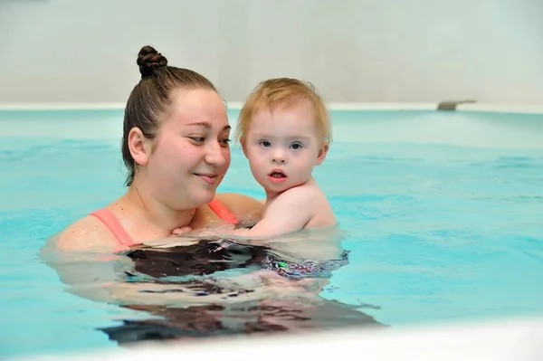 Доросла жінка тримає дитину з синдромом Дауна на руках, стоячи у воді в дитячому басейні з блакитною водою. Вона дивиться на дитину і посміхається. Портрет. Горизонтальна орієнтація . Стокове Фото