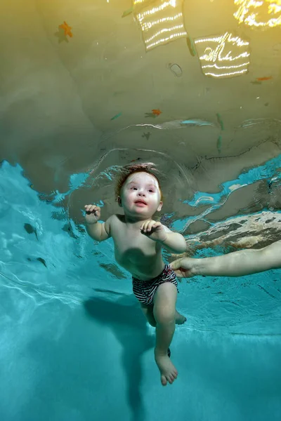 Un niño lindo con síndrome de Down está nadando con los ojos abiertos en la piscina, y su madre lo ayuda y lo apoya con las manos. Un niño con una discapacidad. Retrato. Orientación vertical. — Foto de Stock