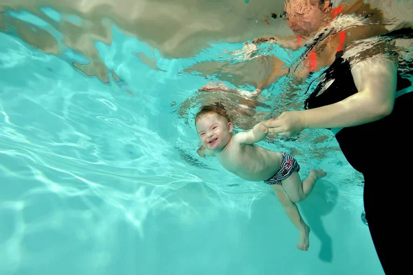 Uma criança rindo com uma deficiência com síndrome de Down nada debaixo d 'água em uma piscina infantil com água azul-turquesa, e sua mãe segura suas mãos. Deficiência infantil. Orientação horizontal. — Fotografia de Stock