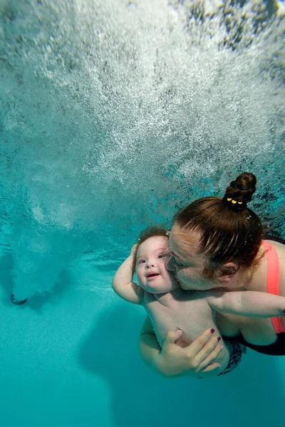 Mamá abraza y besa a su bebé sonriente con síndrome de Down bajo el agua mientras nada en la piscina, y el niño mira los chorros de agua y burbujas. El concepto de felicidad. Orientación vertical. — Foto de Stock