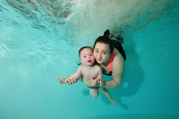 Feliz mamá y el niño con síndrome de Down bucear, nadar y posar para la cámara bajo el agua en una piscina de agua turquesa. Una mujer abraza a un bebé, mira a la cámara y sonríe. Retrato. Orientación horizontal — Foto de Stock