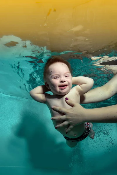 Щасливий маленький хлопчик з синдромом Дауна в руках батьків під водою в бірюзовому басейні. Дитина дивиться в камеру з руками за голову і посміхається. Вертикальна орієнтація . Ліцензійні Стокові Зображення