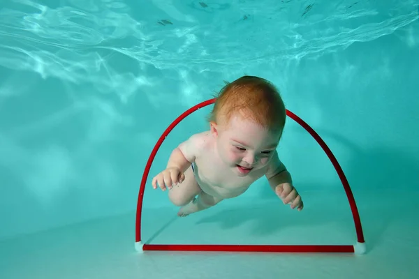다운 증후군 이 있는 귀엽고 작은 아이는 물 속에서 놀다가 수영장 바닥에 있는 훌라후프를 통해 수영을 합니다. 장애가 있는 아이의 행복 한 어린 시절에 대한 개념입니다. 수평 방향. 스톡 사진