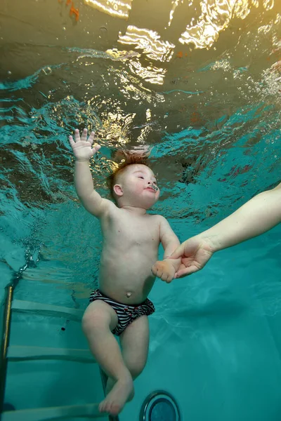 Mamãe segura a mão de um bebê com síndrome de Down, que está nadando debaixo d 'água em uma piscina infantil com água azul. Deficiência infantil. Conceito. Retrato. Orientação vertical. Fotos De Bancos De Imagens Sem Royalties