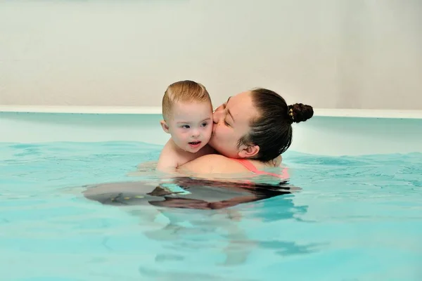 Мати та її дитина з синдромом Дауна плавають та грають у дитячому басейні з блакитною водою. Жінка обіймає і цілує свою дитину з обмеженими можливостями. Портрет. Горизонтальна орієнтація . Стокова Картинка