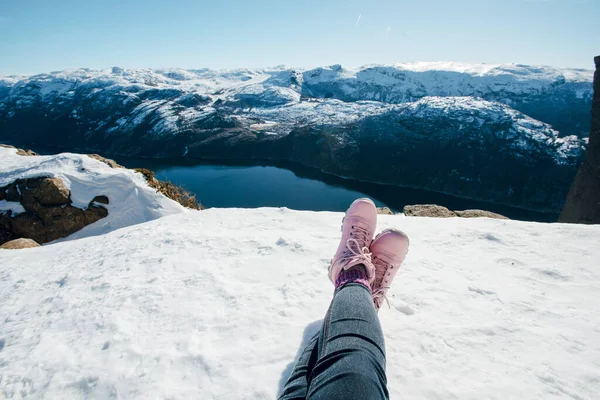 Woman legs in pink boots. Woman sitting on the snow. Scenic landscape of river channel, sea between rocky shore with snow. Top view of the Pulpit Rock, Preikestolen