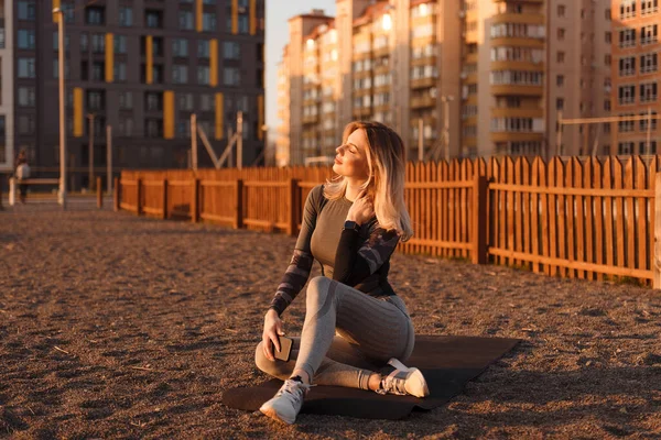 Sporty young woman sitting on yoga mat near playground with closed eyes, holding cellphone in her hand. The girl enjoys the sunset after an active workout.