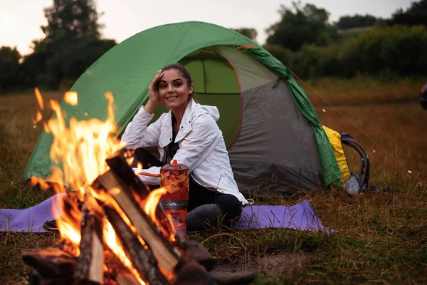 Happy backpacker woman sitting in entrance of tourist tent at campfire and looking away near forest under sky. Camping at the forest concept