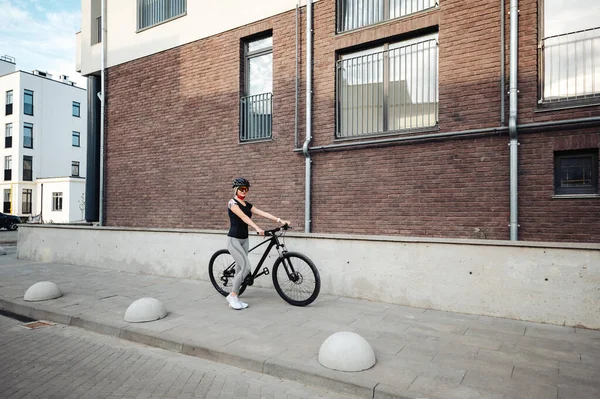 Young caucasian woman with slender body walking outdoors with black bike. Active female blonde wearing sport clothes, safety helmet and mirrored glasses. Cycling concept.