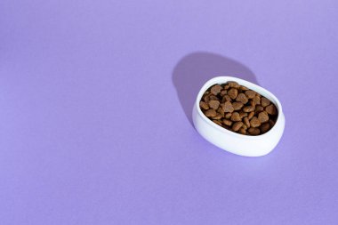 Beyaz kasenin üst görüntüsü, kuru kedi mamasıyla mor, leylak arka plan.