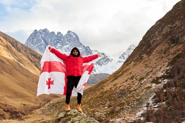 Jovem feliz posando com a bandeira nacional da Geórgia de pé ao ar livre olha para picos cobertos de neve. Homem positivo celebrando o dia da independência da Geórgia — Fotografia de Stock