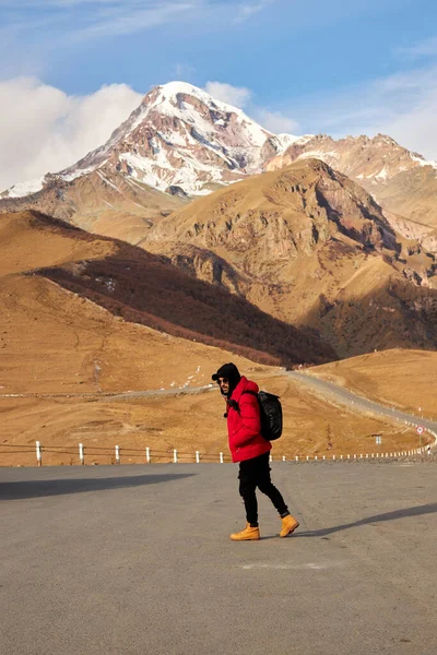 Raveler com mochila caminhadas sozinho na região montanhosa. Backpacker andando pela estrada de encosta com montanha nevada e céu azul no fundo — Fotografia de Stock