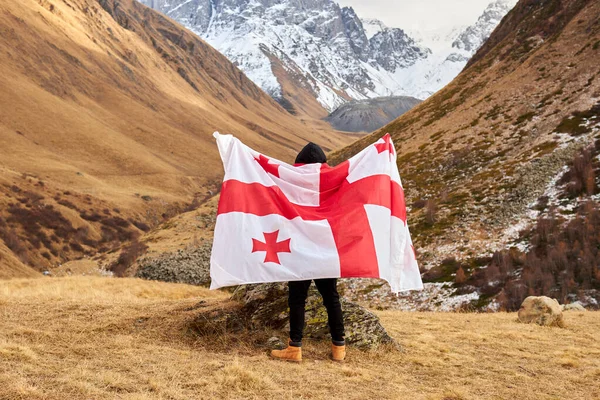 Vista posterior de joven feliz posando con la bandera nacional de Georgia de pie al aire libre mira picos nevados — Foto de Stock