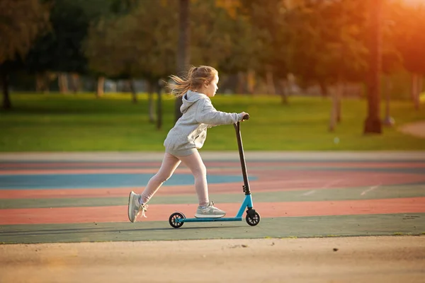 Retrato de una niña pequeña y activa montando scooter en la carretera en el parque al aire libre en el día de verano. Deporte de actividad infantil estacional. Vida saludable de la infancia — Foto de Stock