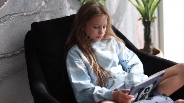 ソファ一人で座っているデジタルタブレット技術デバイスを使用して好奇心旺盛なかわいい子供の女の子 小さな子供は自宅でパッドコンピュータサーフィンインターネットプレイゲームを保持しています 子供技術中毒の概念 — ストック動画