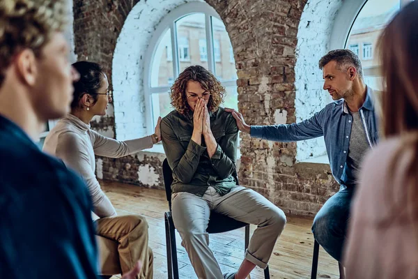 During group therapy sessions participants supporting crying desperate man, provide psychological assistance talking encouraging words share mental pain try to help, struggle with addictions treatment. Selective focus