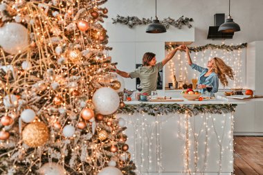 Ön planda Noel ağacı olan sevimli bir çift mutfakta dans ediyor. Birkaç tanesine odaklan.