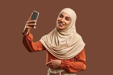Bej tesettürlü güzel Müslüman kadın elinde bir telefon tutuyor, gülümsüyor ve kahverengi bir arka planın yanında selfie çekiyor..