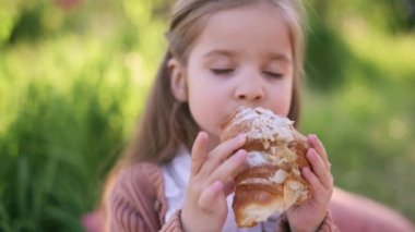 Küçük tatlı kız iştah açıcı kruvasan yiyor ve gülümseyen kameraya bakıyor. Açık hava portresi..
