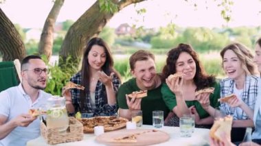 Bir grup arkadaş dışarıda birlikte dinleniyorlar. Gençler masada oturmuş, sohbet ediyor ve pizza yiyorlar..