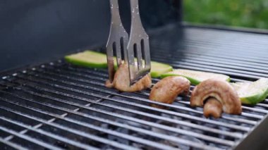 Vegetables are grilled. Barbecue of mushrooms and zucchini, close-up. The cook puts the champignons on the grill grate with tongs.