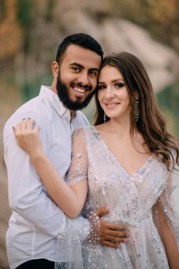 Irklar arası ilişkiler. Orta Doğulu damat ve beyaz gelin güzel bir göle karşı sarılıyor. Portreyi kapat.