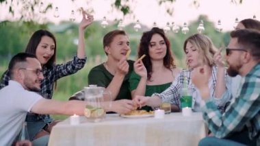 Bir grup arkadaş dışarıda birlikte dinleniyorlar. Gençler masada oturmuş, eğleniyor, konuşuyor ve yemek yiyorlar..