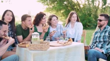 Bir grup arkadaş dışarıda birlikte dinleniyorlar. Gençler masada oturmuş, sohbet ediyor ve pizza yiyorlar..