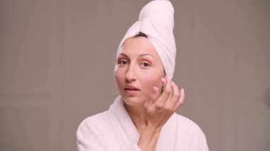 Young attractive woman in a bathrobe applies cream on her face in the bathroom. Close-up of the face.