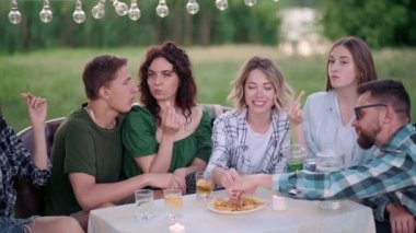 Bir grup arkadaş dışarıda birlikte dinleniyorlar. Gençler masada oturmuş, eğleniyor, konuşuyor ve yemek yiyorlar..