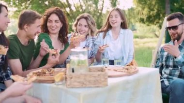 Bir grup arkadaş dışarıda birlikte dinleniyorlar. Gençler masada oturmuş, sohbet ediyor ve pizza yiyorlar..