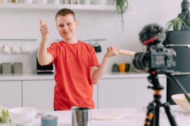 A child blogger makes a tutorial video at home. Joyful boy shows a thumbs up at the camera.