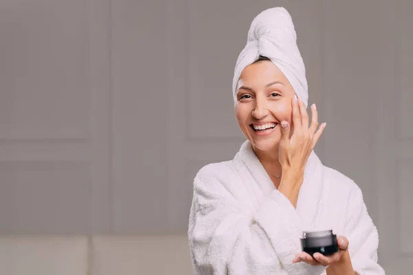 Young smiling attractive woman in a bathrobe applies cream on her face in the bathroom.