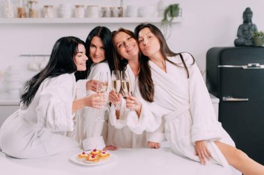 A group of women spend leisure time. Young attractive females in bathrobes drink champagne and chat merrily at home.