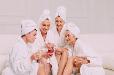A group of women spend leisure time. Four Young attractive women in bathrobes look at the smartphone screen and smile.