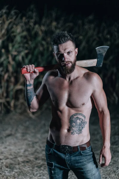 Scary man with ax stands in corn field and looks into camera at night. Halloween concept