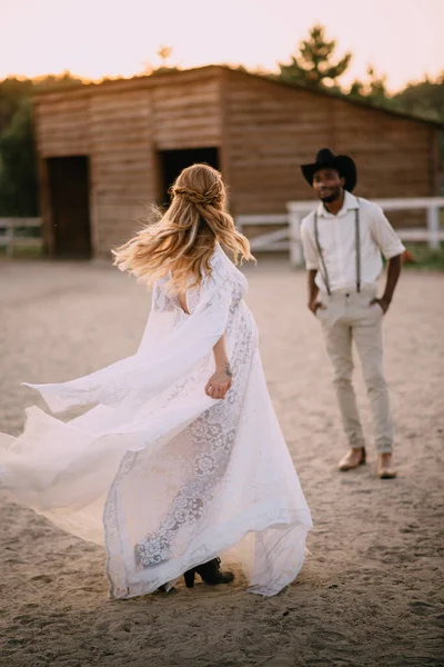 Cowboy style wedding. Caucasian bride in boho style dress dances in front of her African groom on ranch.