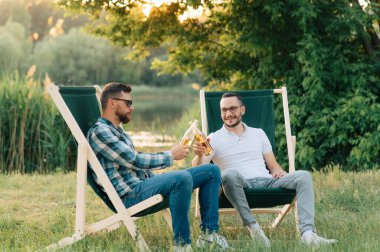 Kamp sandalyelerinde oturup bira içip dışarıda sohbet eden iki adam..
