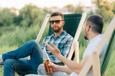 Kamp sandalyelerinde oturup bira içip dışarıda sohbet eden iki adam..