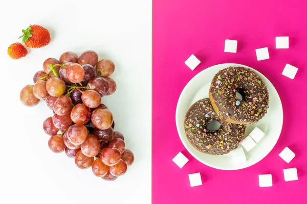 Healthy and unhealthy food, flat layer, two-color background, chocolate donuts with sprinkles on a fuchsia background, juicy delicious grapes and strawberries on white, natural sugar vs harmful processed