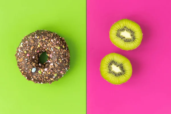 Healthy and unhealthy food, flat layer, two-color chocolate donuts with sprinkles on a green background vs juicy kiwi on a fuchsia background, natural and harmful processed sugar