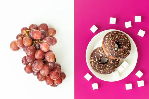 Healthy and unhealthy food, flat layer, two color background, chocolate donuts with sprinkles on a fuchsia background, juicy delicious grapes on white, Natural sugar vs harmful processed