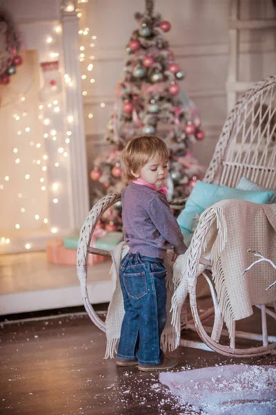 A boy with a pink tie stands near a wheelchair on the background of a Christmas tree and a wall decorated with garlands