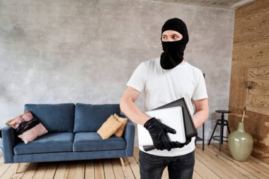 Siyah kar maskeli bir hırsız dairesinden modern ve pahalı bir laptop çalıyor. Maskeli yüz. Hırsız evden televizyon çalıyor.