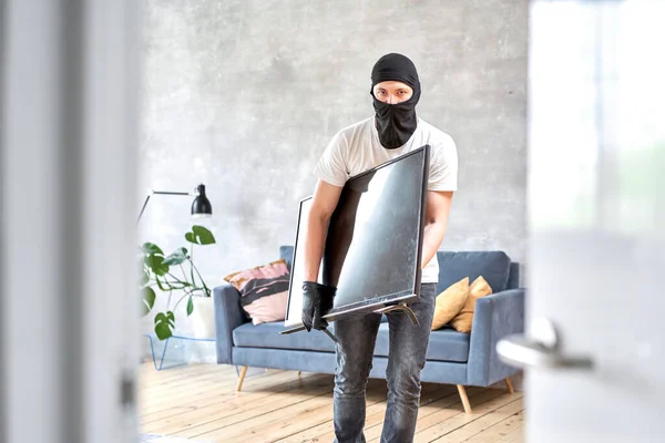 Tjuv med svart balaklava stjäl modern dyr tv. Maskerat ansikte. Man inbrottstjuv stjäla tv från huset — Stockfoto